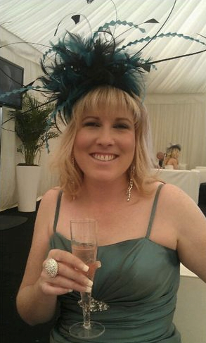 a picture of charley at a wedding in a big blue fascinator and with champagne in hand. She's smiling.